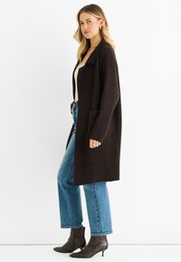 Woman wearing long dark brown cardigan, light beige top, blue jeans, and dark patterned ankle boots, standing against a white background.