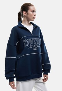 Granatowy sweter z ćwierć zamkiem, z białymi akcentami i napisem "NEW YORK The Bronx 2002" z przodu. Wykonany z miękkiego, ciepłego materiału.