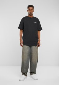 Sort bomulds oversized t-shirt med logo på brystet, parret med løstsiddende lysegrønt denim og hvide sneakers. Enkelt baggrund.