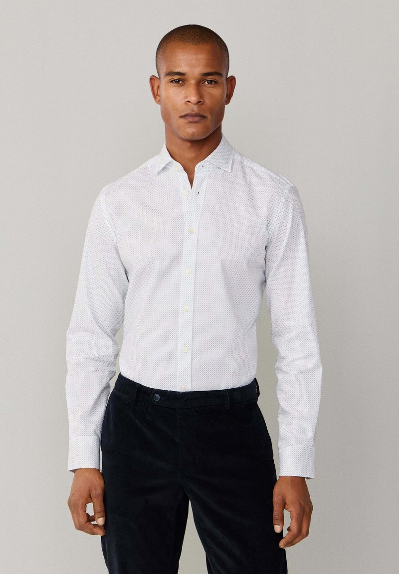 Witte overhemdd met een subtiel stippenpatroon, lange mouwen en een puntige kraag, gecombineerd met donker corduroy broek.