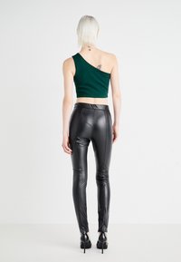 Person med kort vitt hår bär en mörkgrön enskuldrad crop top, svarta läderbyxor och svarta högklackade skor, stående mot en vit bakgrund.
