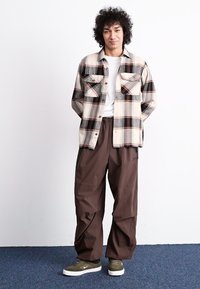 Pleds vīriešu krekls bēšā, brūnā un pelēkā krāsā; apvienots ar brīviem brūniem biksēm un olīvzaļiem kedām ar baltām zālēm.