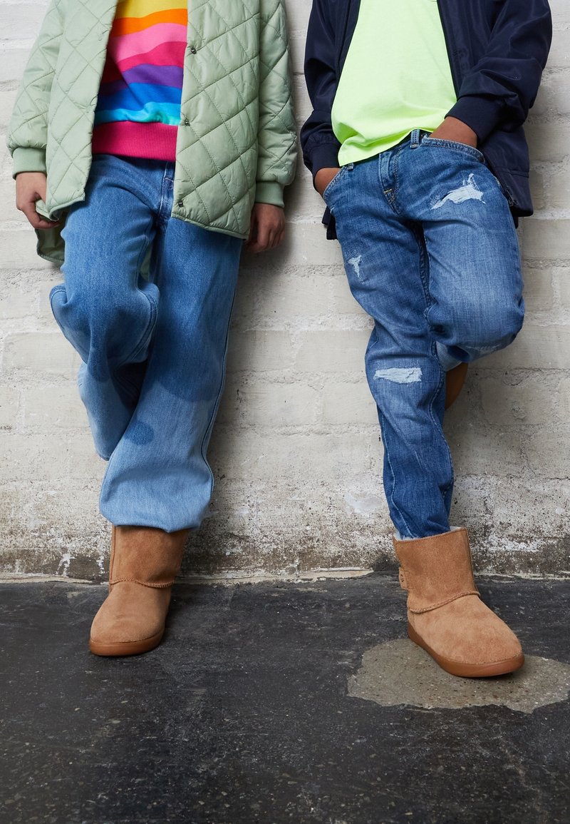 Deux personnes appuyées contre un mur en briques blanches, portant des jeans, des bottes beige et des vestes décontractées avec des chemises colorées en dessous.