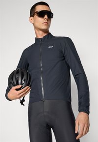 Black cycling jacket with a high collar and zip front, paired with black padded cycling shorts, and a black helmet in the model's hand.