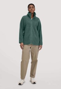 Green fleece jacket with high collar and full zip, paired with beige cargo pants. The texture is soft, with subtle seam detailing and side pockets.