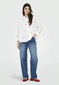 Mujer joven de pie con la mano en el bolsillo, vistiendo una camisa blanca con estampado de cerezas rojas, jeans azules y zapatos de tacón rojos.