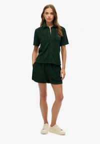 Camisa de botones de manga corta de color verde oscuro con un bolsillo, combinada con pantalones cortos a juego. La tela presenta un suave patrón texturizado. Zapatillas de color claro.