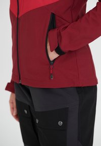 Röd och svart jacka med en zippered ficka, med en texturerad tyg och kontrasterande färger. Ärmarna har justerbara manschetter med kardborre.