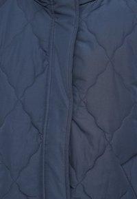 Μπλε ναυτικό μπουφάν με καπιτονέ ύφασμα, ομαλή υφή, διακοσμημένο με διαμάντι σχέδια και εμπρόσθιο κλείσιμο με φερμουάρ.