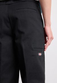 Detailní záběr na osobu v černých pracovních kalhotách s zadními a bočními kapsami, ukazující malou barevnou značkovou nášivku na boční kapse.