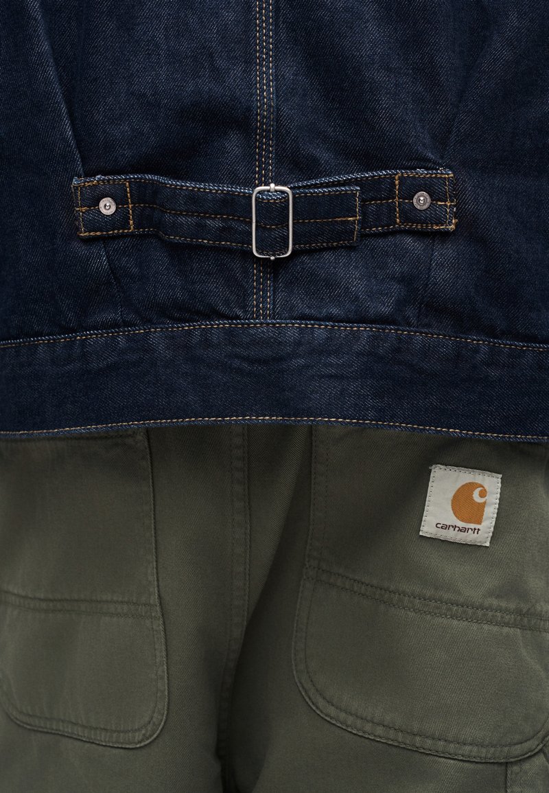 Nahaufnahme eines verstellbaren Rückengurtes einer Jeansjacke über olivgrünen Hosen mit Carhartt-Logopatch auf der rechten Tasche.