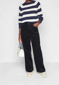 Pull rayé bleu marine et blanc, texture côtelée. Pantalons wide-leg en velours côtelé noir. Modèle tient un sac blanc avec des poignées bleues. Baskets beiges.