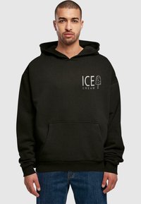 Černá bavlněná mikina s kapucí, přední klokankou, nastavitelnou kapucí a grafikou "ICE CREAM" s designem nanuku. Nosená s modrými džínami.