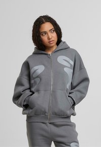 Sudadera gris con cremallera hecha de una tela suave. Presenta un gran logotipo gráfico en gris más claro, bolsillos frontales y puños y dobladillo acanalados.