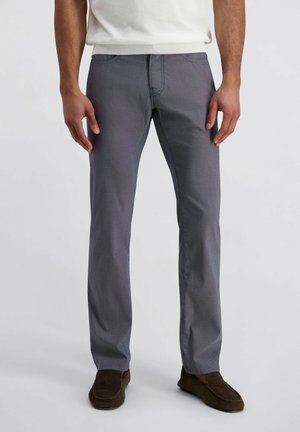 Mann trägt graue Hosen mit geradem Bein, braune Loafer und ein weißes Hemd, steht vor einem schlichten hellen Hintergrund.