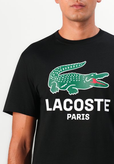 Черна памучна тениска с изображение на зелена крокодилска емблема и "LACOSTE PARIS" с удебелен бял текст. Къси ръкави и кръгло деколте.