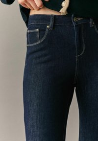Jeans de denim azul oscuro con corte ajustado, que presentan un distintivo pespunte amarillo y un pequeño bolsillo frontal. La cinturilla cuenta con un cierre de botón.