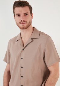 Kortærmet, lysebrun skjorte med knapper lavet af glat stof. Har krave, sorte knapper og en afslappet pasform.