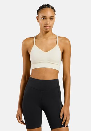 Femme aux cheveux tressés portant un brassière de sport beige clair et un short cycliste noir taille haute, debout devant un fond uni.