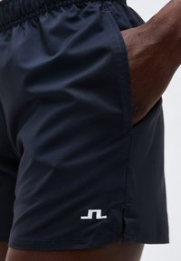 Κοντινό πλάνο ενός ατόμου που φοράει μαύρο αθλητικό σορτς με ένα μικρό λευκό λογότυπο κοντά στο τελείωμα και το χέρι ακουμπισμένο μέσα στην τσέπη.