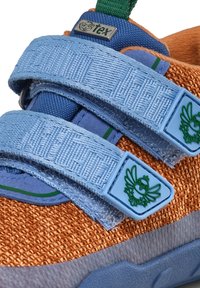 Kinderturnschuhe aus orangefarbenem, gewebtem Stoff, mit blauen Klettverschlüssen und grünen Akzenten. Enthält ein blaues Logodetail am Riemen. Leichtes Design.