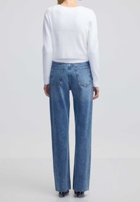 Femme portant un jean droit taille haute bleu clair et un pull blanc à manches longues, debout de dos sur un fond uni.