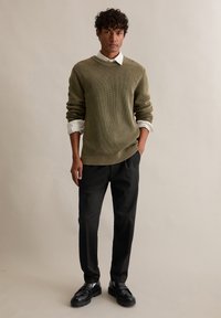 Olivově zelený svetr s žebrovaným vzorem a kulatým výstřihem, vrstvený přes bílou košili s límečkem, doplněný černými upravenými kalhotami a černými mokasínami.