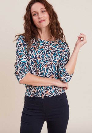 Bluse mit Blumenmuster in Blau, Weiß und Orange; verfügt über Dreiviertelärmel und einen gerafften Saum. Getragen mit dunklen Jeans.