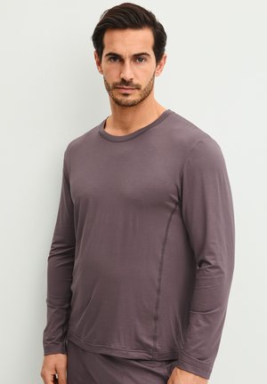 Man met donker haar en baard, dragend een nauwsluitend bruin shirt met lange mouwen, staand tegen een eenvoudige lichte muur.