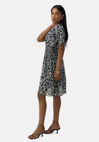 Vestido con estampado floral sobre un fondo oscuro, de mangas cortas, cintura elástica y una silueta fluida. Lleva sandalias negras de tiras.