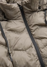 Beige pufferjack met een gequilt ontwerp, zwarte ritsen, verstelbare trekkoorden en een gladde, glanzende textuur.