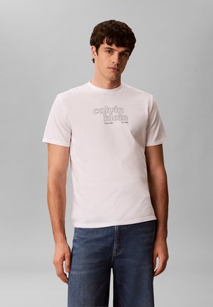 Jonge man draagt een wit Calvin Klein T-shirt met logo en blauwe spijkerbroek, staand tegen een effen lichtgrijze achtergrond.