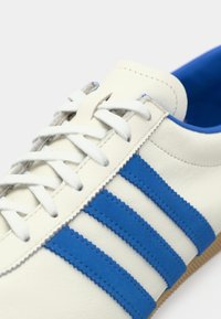 Λευκό αθλητικό παπούτσι με μπλε σουέντ ρίγες, κλωστές υφάσματος, λαστιχένια σόλα και μπλε εσωτερική επένδυση. Διαθέτει ανάγλυφο δερμάτινο επάνω μέρος.