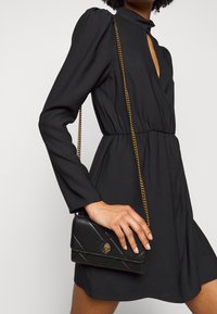 Woman wearing a black long-sleeve dress with puffed shoulders, holding a small black quilted handbag with a gold chain strap and clasp.