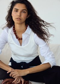 Jeune femme aux longs cheveux bouclés portant une chemise blanche et un débardeur, collier marron en perles, assise les mains jointes et pull noir noué à la taille.