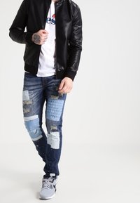 Giubbotto bomber in pelle nera sopra una t-shirt bianca, abbinato a jeans blu strappati con toppe azzurre chiare. Sneakers grigie completano il look.