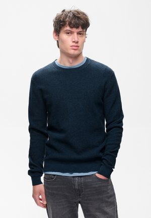 Ung mand med krøllet brunt hår iført en mørkeblå sweater og grå jeans, stående med den ene hånd i lommen mod en ensfarvet baggrund.