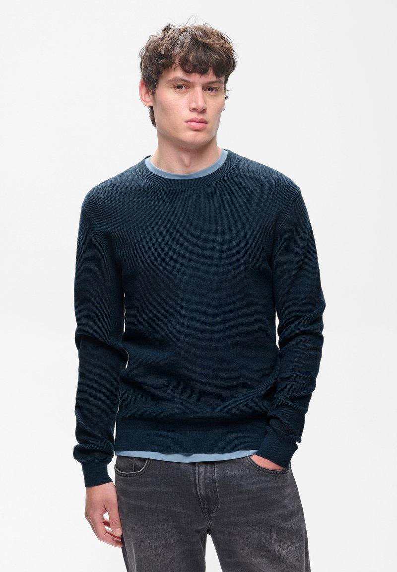 Jeune homme aux cheveux châtains bouclés portant un pull bleu foncé et un jean gris, debout avec une main dans la poche devant un fond uni.