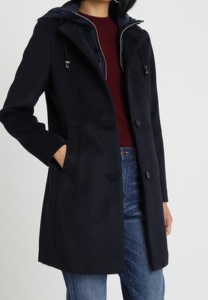 Vrouw draagt een marineblauwe gewatteerde jas met capuchon over een bordeauxrode trui en een blauwe spijkerbroek, met één hand in de jaszak.