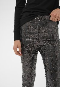 Črne bleščeče hlače z oprijetim krojem, ki imajo teksturirano površino in sijoč učinek zaradi bleščic razporejenih v vertikalne črte.