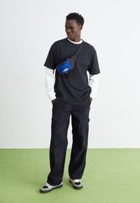 Black T-shirt layered over a white long-sleeve, paired with dark patterned cargo pants and a blue crossbody bag. Textured black sneakers.