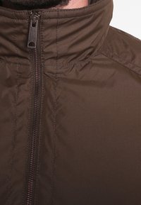 Veste isolante marron avec un col haut, dotée d'une fermeture éclair sur toute la longueur et d'une texture matelassée, fabriquée en matériau lisse et résistant à l'eau.
