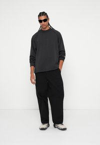 Liela izmēra melns sporta krekls ar raglanu piedurknēm, apvienots ar melnajām kravas biksēm un gaišajiem pelēkajiem trenažieriem. Saulesbrilles kā ievērojams aksesuārs.