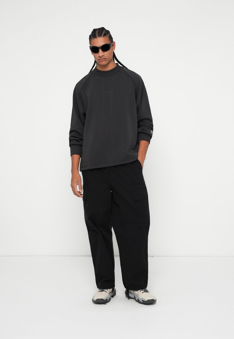 Übergröße schwarzer Pullover mit Raglanärmeln, kombiniert mit schwarzen Cargo-Hosen und hellgrauen Sneakern. Sonnenbrille als auffälliges Accessoire.