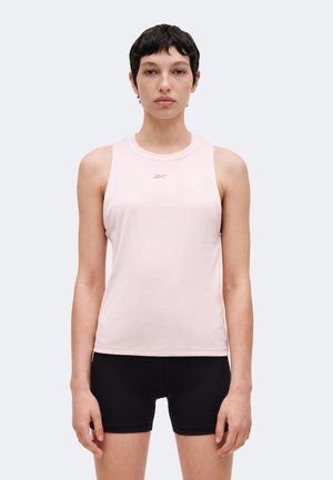 Jeune personne aux cheveux courts et foncés portant un haut de sport sans manches rose clair et un cuissard de cyclisme noir, debout face à l'avant.