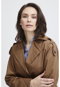 Brauner Trenchcoat mit Reverskragen, Knopfakzenten und strukturiertem Stoff. Darunter ist ein blusiges blaues Shirt sichtbar.