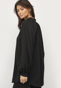 Sort bluse med lange ærmer og struktureret finish, der har en høj hals og elastiske manchetter, hvilket skaber en løs, flydende silhuet.