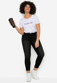Jeans negros ajustados, camiseta corta blanca con el texto "Push Up", cinturón negro, sosteniendo una taza negra, combinados con zapatillas blancas.