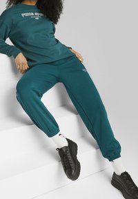 Sudadera de forro polar en color teal con el logo "Puma Sports", combinada con joggers a juego que tienen puños elásticos, y zapatillas negras con suelas texturizadas.