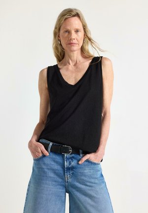 Kvinde med langt blondt hår iført en sort ærmeløs top og blå jeans, stående med hænderne i lommerne foran en ensfarvet baggrund.